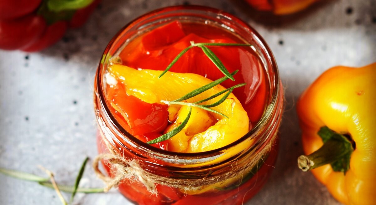 Eingelegte Paprika in einem Glas auf einem grauen Steintisch mit Kräutern und Olivenöl. Alles von schräg oben fotografiert.