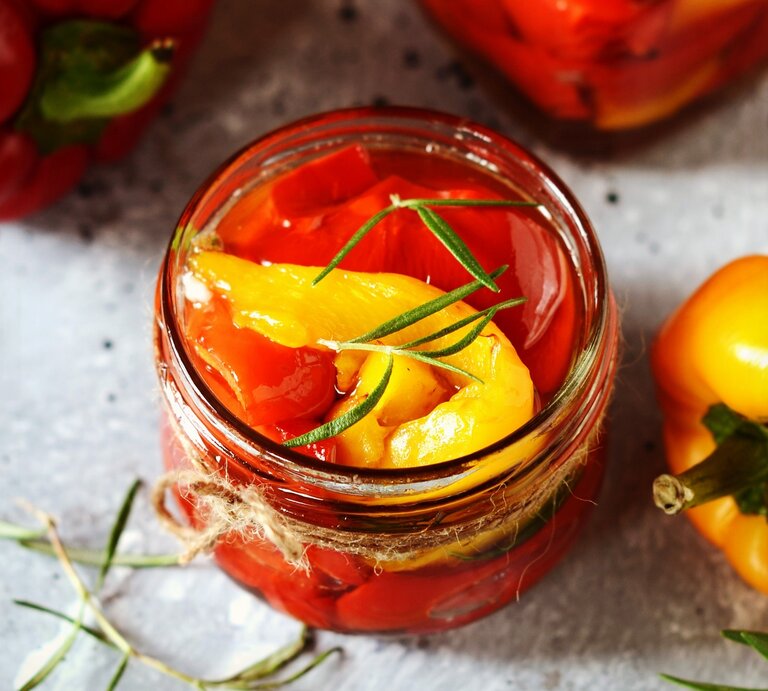 Eingelegte Paprika in einem Glas auf einem grauen Steintisch mit Kräutern und Olivenöl. Alles von schräg oben fotografiert. | © Shutterstock/ teleginatania