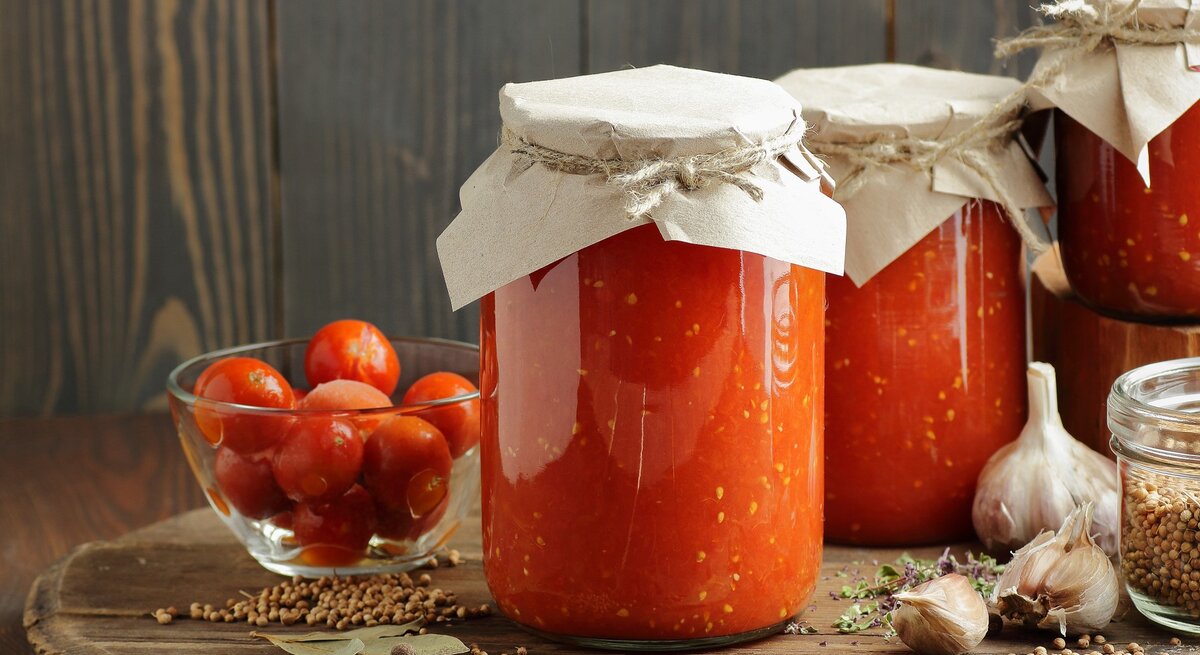 Mehrere Gläser mit eingekochter Tomate auf einem dunklen Holztisch mit Gewürzen und frischen Kirschtomaten von der Seite fotografiert.