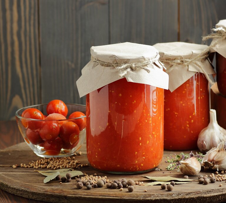 Mehrere Gläser mit eingekochter Tomate auf einem dunklen Holztisch mit Gewürzen und frischen Kirschtomaten von der Seite fotografiert. | © Shutterstock/ teatian