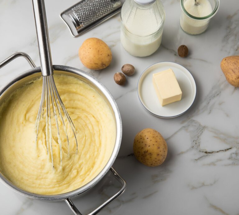 Kartoffelpüree in einem Topf mit Schneebesen, daneben Kartoffeln, Butter, Milch und Muskatnüsse auf hellem Marmortisch. | © AdobeStock/ Christin Klose