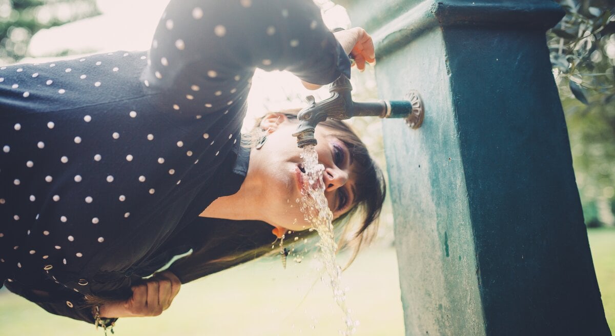 Nahaufnahme einer Frau in gepunkteter Bluse, die sich über einen Wasserhahn im Park beugt und trinkt.