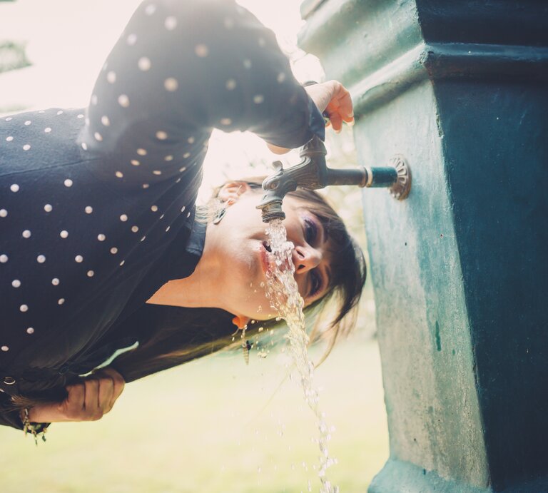 Nahaufnahme einer Frau in gepunkteter Bluse, die sich über einen Wasserhahn im Park beugt und trinkt. | © Getty Images/kaisersosa67