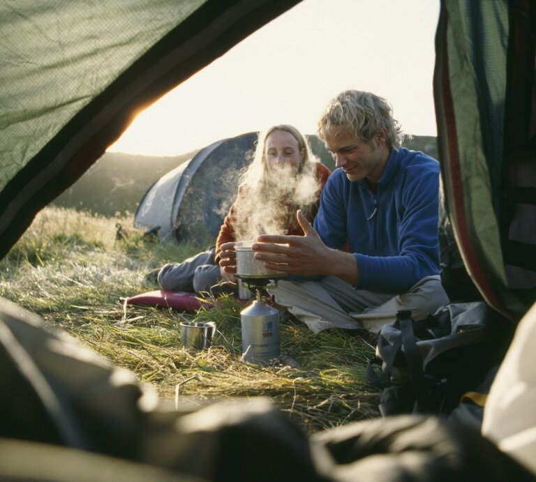 Ein Paar beim Zelten sitzt vor einem Gaskocher, auf dem etwas heiß aufkocht. Im Hintergrund geht die Sonne auf.  | © Getty Images/Ted Levine