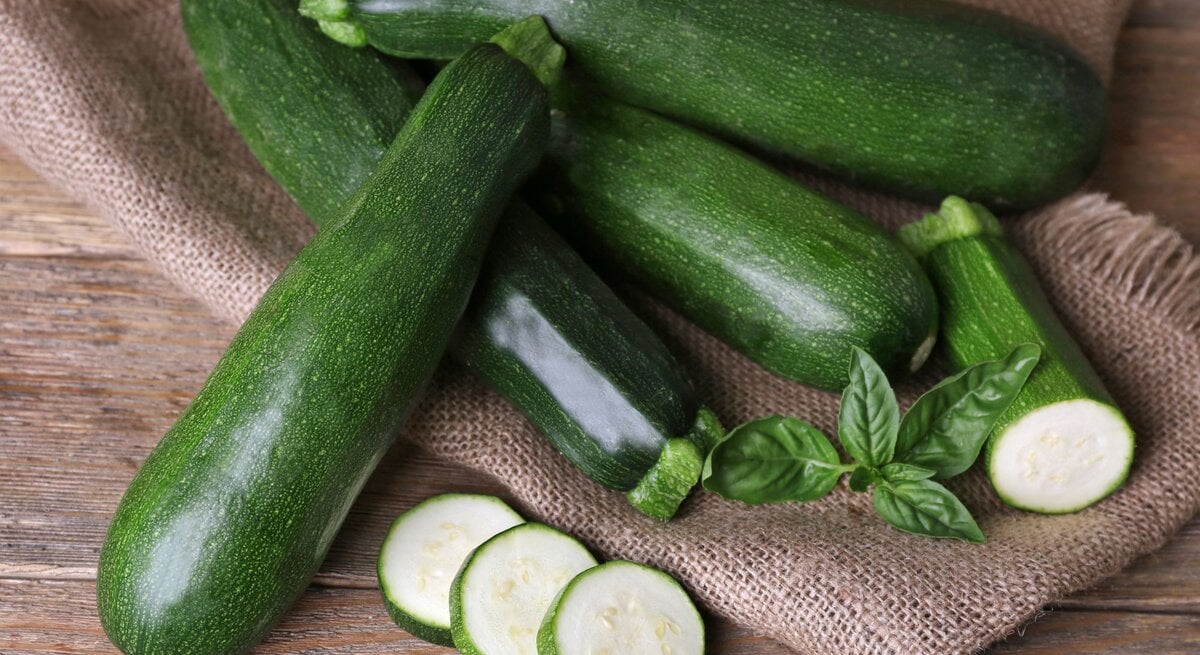 Frische grüne Zucchini liegen auf einem groben Jutesack, mit geschnittenen Scheiben und Basilikumblättern dekoriert.