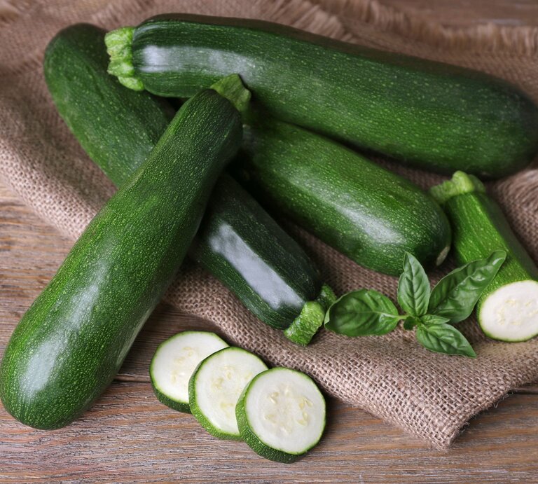 Frische grüne Zucchini liegen auf einem groben Jutesack, mit geschnittenen Scheiben und Basilikumblättern dekoriert. | © Shutterstock/Africa Studio