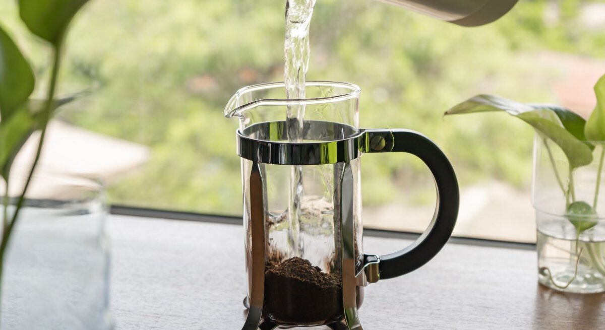 Wasser wird aus einem grauen Kessel in eine French Press mit Kaffee gegossen, neben grünen Pflanzen.