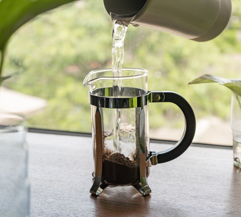 Wasser wird aus einem grauen Kessel in eine French Press mit Kaffee gegossen, neben grünen Pflanzen. | © AdobeStock/wachiwit