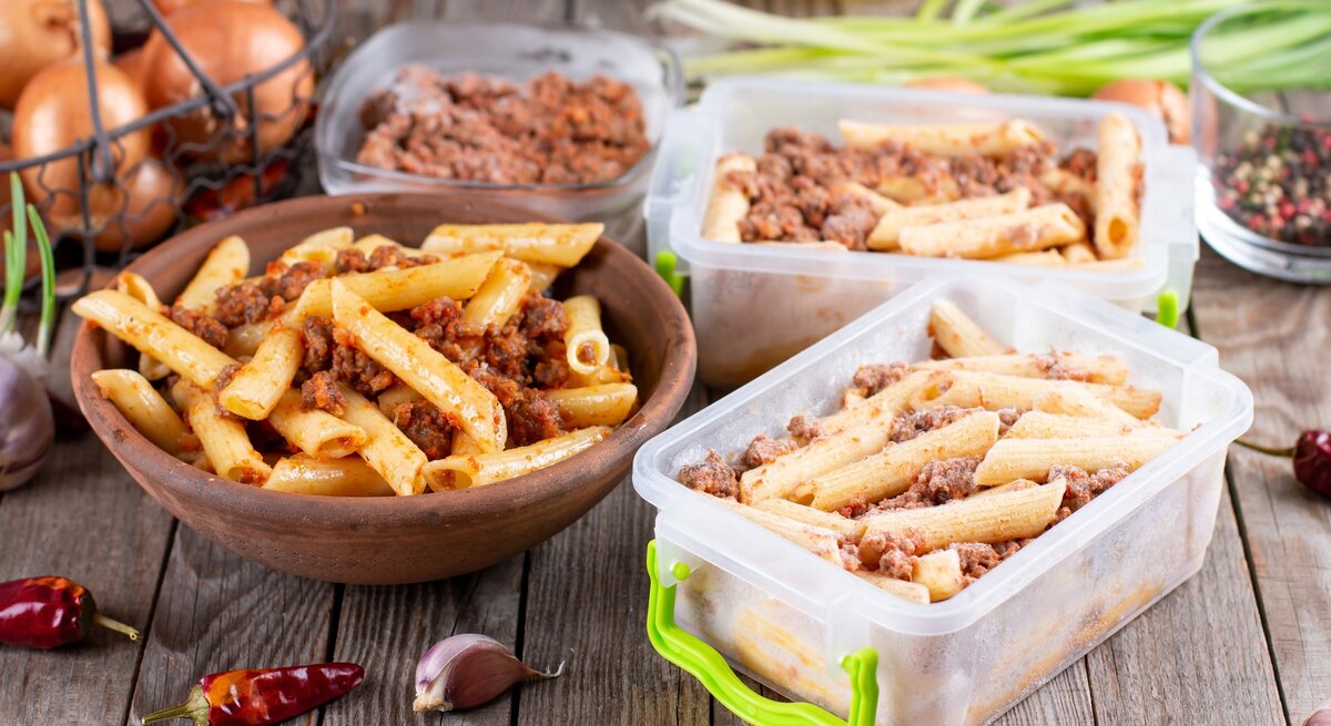 Das Bild zeigt eine hölzerne Schüssel gefüllt mit Pasta, die anscheinend mit einer Bolognese-Sauce gemischt ist, sowie mehrere Behälter mit der gleichen Pasta, die für die Lagerung oder das Einfrieren vorbereitet zu sein scheinen. Im Hintergrund sind einige Küchenzutaten und -utensilien sichtbar, darunter Zwiebeln, Knoblauch, Pfefferkörner, grüne Zwiebeln und Olivenöl, was auf eine frische Zubereitung der Mahlzeiten hinweist. Die Szene deutet auf das Konzept des Meal-Prepping hin, bei dem Mahlzeiten im Voraus zubereitet und dann für später gelagert werden.