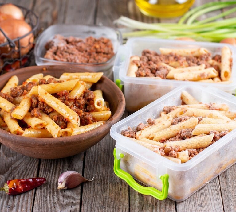 Das Bild zeigt eine hölzerne Schüssel gefüllt mit Pasta, die anscheinend mit einer Bolognese-Sauce gemischt ist, sowie mehrere Behälter mit der gleichen Pasta, die für die Lagerung oder das Einfrieren vorbereitet zu sein scheinen. Im Hintergrund sind einige Küchenzutaten und -utensilien sichtbar, darunter Zwiebeln, Knoblauch, Pfefferkörner, grüne Zwiebeln und Olivenöl, was auf eine frische Zubereitung der Mahlzeiten hinweist. Die Szene deutet auf das Konzept des Meal-Prepping hin, bei dem Mahlzeiten im Voraus zubereitet und dann für später gelagert werden. | © Getty Images/Qwart