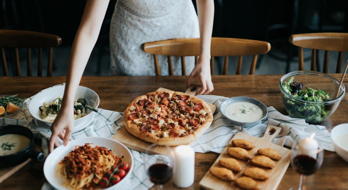 Eine Person in einem weißen Spitzenkleid bereitet am Tisch ein italienisches Essen vor. Auf dem Tisch befindet sich eine Vielfalt an Gerichten: eine Pizza mit reichlich Belag auf einem Holzbrett, ein Teller mit Spaghetti und Tomatensoße, ein Salat in einer Glaschüssel, eine Schüssel mit einer hellen, cremigen Suppe und eine Platte mit goldbraunen, gebackenen Teigtaschen. Zwei Gläser Rotwein und eine Kerze tragen zur gemütlichen Atmosphäre bei.