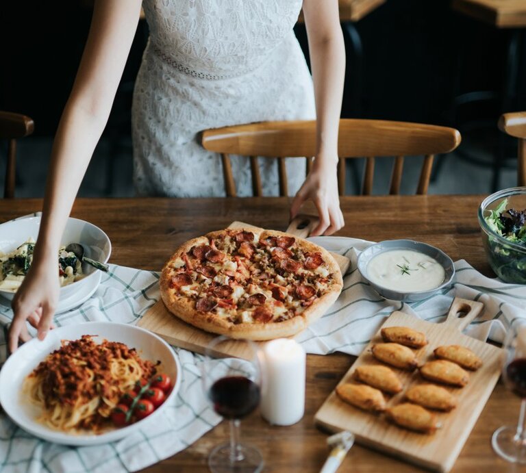 Eine Person in einem weißen Spitzenkleid bereitet am Tisch ein italienisches Essen vor. Auf dem Tisch befindet sich eine Vielfalt an Gerichten: eine Pizza mit reichlich Belag auf einem Holzbrett, ein Teller mit Spaghetti und Tomatensoße, ein Salat in einer Glaschüssel, eine Schüssel mit einer hellen, cremigen Suppe und eine Platte mit goldbraunen, gebackenen Teigtaschen. Zwei Gläser Rotwein und eine Kerze tragen zur gemütlichen Atmosphäre bei. | © GettyImages/AsiaVision