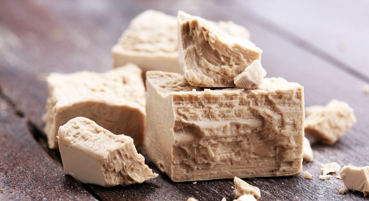 Mehrere Stücke frischer Hefe liegen auf einer dunklen Holzoberfläche. Die Hefe hat eine charakteristisch poröse Textur mit einer cremigen Farbe. Einige Bröckchen liegen verstreut um die größeren Blöcke herum, was ihre weiche und krümelige Beschaffenheit hervorhebt. Das Bild vermittelt einen Eindruck von der Backzutat in ihrer natürlichen, unverarbeiteten Form.