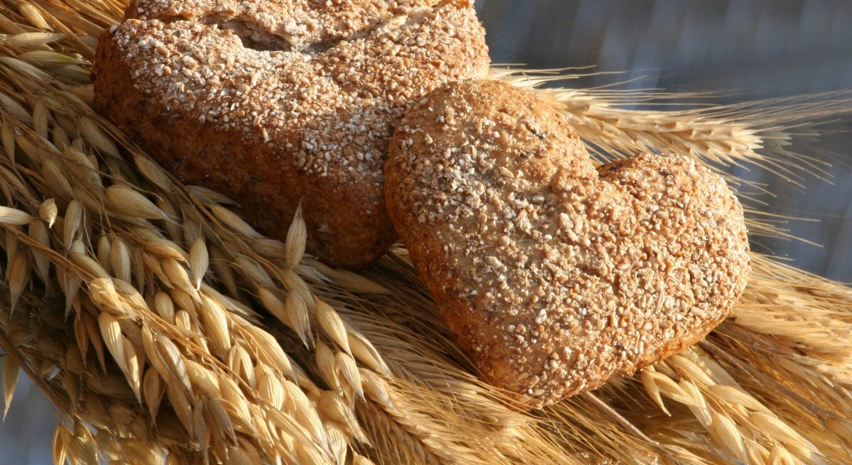 Zwei herzförmige Vollkornbrötchen, bestreut mit Sesamkörnern, liegen auf einem Bündel goldener Weizenähren. Die warme Beleuchtung betont die goldbraune Farbe des Gebäcks und das strahlende Gelb des Weizens, was eine Verbindung von frisch gebackenem Brot und seinem Ursprung im Kornfeld suggeriert. Die Komposition strahlt eine rustikale und gemütliche Atmosphäre aus.