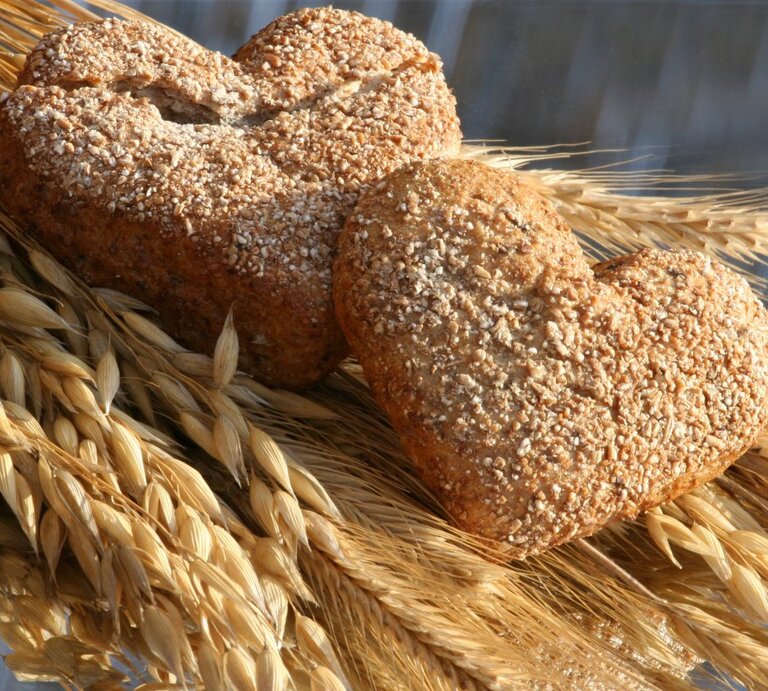 Zwei herzförmige Vollkornbrötchen, bestreut mit Sesamkörnern, liegen auf einem Bündel goldener Weizenähren. Die warme Beleuchtung betont die goldbraune Farbe des Gebäcks und das strahlende Gelb des Weizens, was eine Verbindung von frisch gebackenem Brot und seinem Ursprung im Kornfeld suggeriert. Die Komposition strahlt eine rustikale und gemütliche Atmosphäre aus. | ©  iStock/diephosi