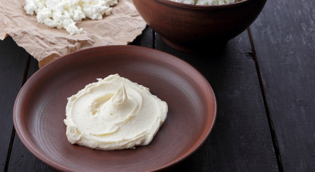 Ein Schlag frischer Quark oder Topfen ist kunstvoll auf einem dunkelbraunen Keramikteller serviert, der auf einer schwarzen Holzoberfläche steht. Im Hintergrund, außerhalb des Fokus, ist eine größere Menge desselben Produkts zu sehen, die in einer dunkelbraunen Keramikschüssel auf bewährtem Packpapier präsentiert wird. Die Farben und Texturen unterstreichen die Einfachheit und Natürlichkeit des Lebensmittels