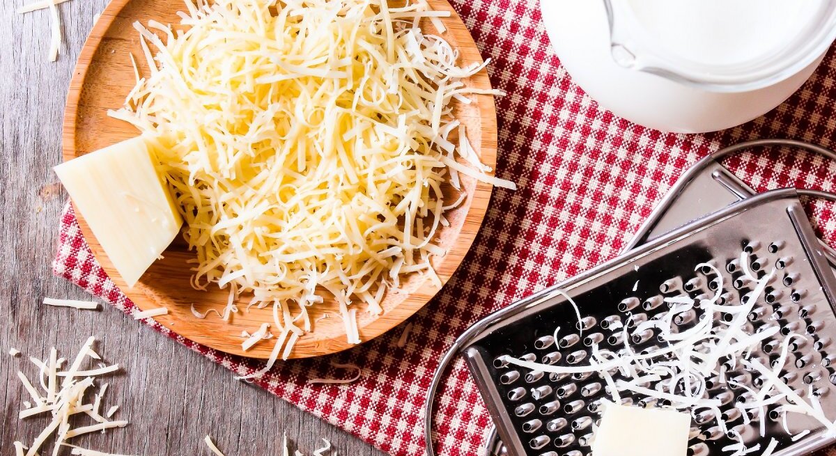 Auf einem rustikalen Holztisch liegt eine runde Holzplatte, auf der frisch geriebener Käse zu einem Haufen aufgetürmt ist. Neben der Platte liegt ein Stück Käse, von dem offensichtlich gerieben wurde. 
