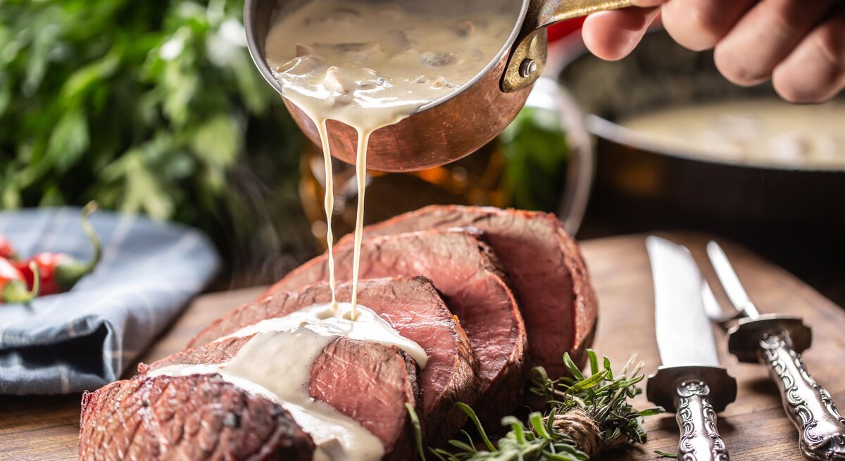 Eine Hand gießt cremige Sauce aus einem kleinen, antiken Kupfertopf über ein saftig aussehendes Stück rosa gebratenes Rindfleisch, das auf einem Holzbrett angerichtet ist. Neben dem Fleisch liegen frische Rosmarinzweige und silberne Besteckteile mit verzierten Griffen. 