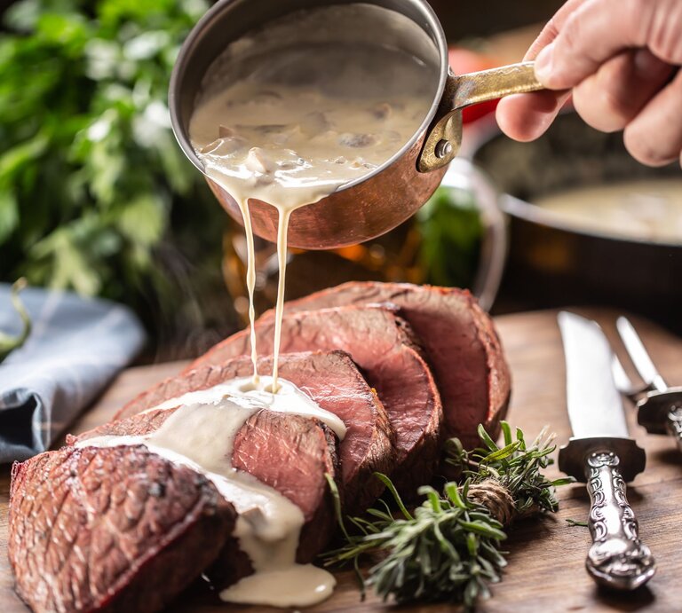 Eine Hand gießt cremige Sauce aus einem kleinen, antiken Kupfertopf über ein saftig aussehendes Stück rosa gebratenes Rindfleisch, das auf einem Holzbrett angerichtet ist. Neben dem Fleisch liegen frische Rosmarinzweige und silberne Besteckteile mit verzierten Griffen.  | © AdobeStock/weyo