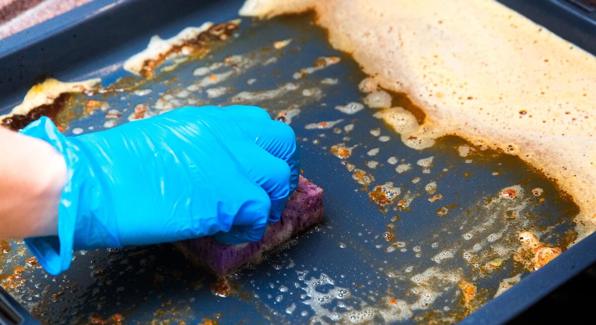 Eine Hand mit blauen Gummihandschuhen schrubbt mit einem Schwamm ein schmutziges Backblech mit Seifenlauge.