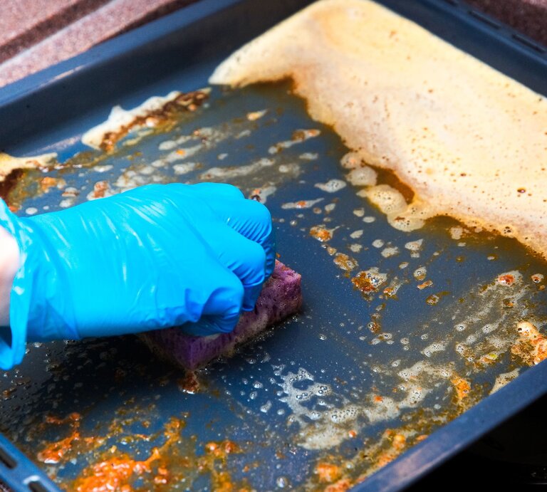 Eine Hand mit blauen Gummihandschuhen schrubbt mit einem Schwamm ein schmutziges Backblech mit Seifenlauge. | © Adobe Stock/ Евгения Маслова