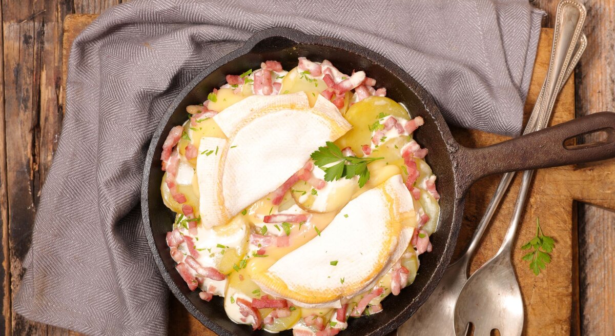 Das Bild zeigt eine Tartiflette, ein traditionelles französisches Gericht aus der Region Savoyen, das in einer gusseisernen Pfanne serviert wird. Es besteht aus Schichten von Kartoffeln, Speck und Zwiebeln, die reichlich mit Reblochon-Käse überbacken sind. Die oberste Käseschicht ist leicht gebräunt und geschmolzen, was auf eine kürzliche Zubereitung im Ofen hinweist. Garniert ist das Gericht mit frischen Kräutern, vermutlich Petersilie, die einen Farbtupfer geben. Das rustikale Holzbrett unter der Pfanne sowie das graue Stofftuch und das antike Besteck runden das Bild eines herzhaften, ländlichen Gerichts ab.