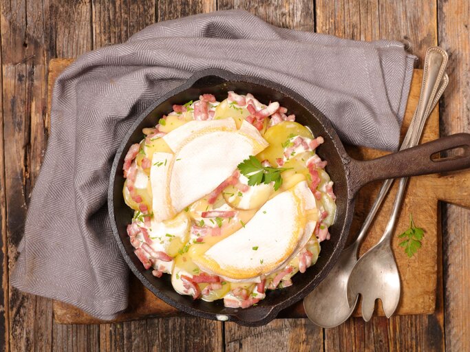 Das Bild zeigt eine Tartiflette, ein traditionelles französisches Gericht aus der Region Savoyen, das in einer gusseisernen Pfanne serviert wird. Es besteht aus Schichten von Kartoffeln, Speck und Zwiebeln, die reichlich mit Reblochon-Käse überbacken sind. Die oberste Käseschicht ist leicht gebräunt und geschmolzen, was auf eine kürzliche Zubereitung im Ofen hinweist. Garniert ist das Gericht mit frischen Kräutern, vermutlich Petersilie, die einen Farbtupfer geben. Das rustikale Holzbrett unter der Pfanne sowie das graue Stofftuch und das antike Besteck runden das Bild eines herzhaften, ländlichen Gerichts ab. | © AdobeStock/	margouillatphotos