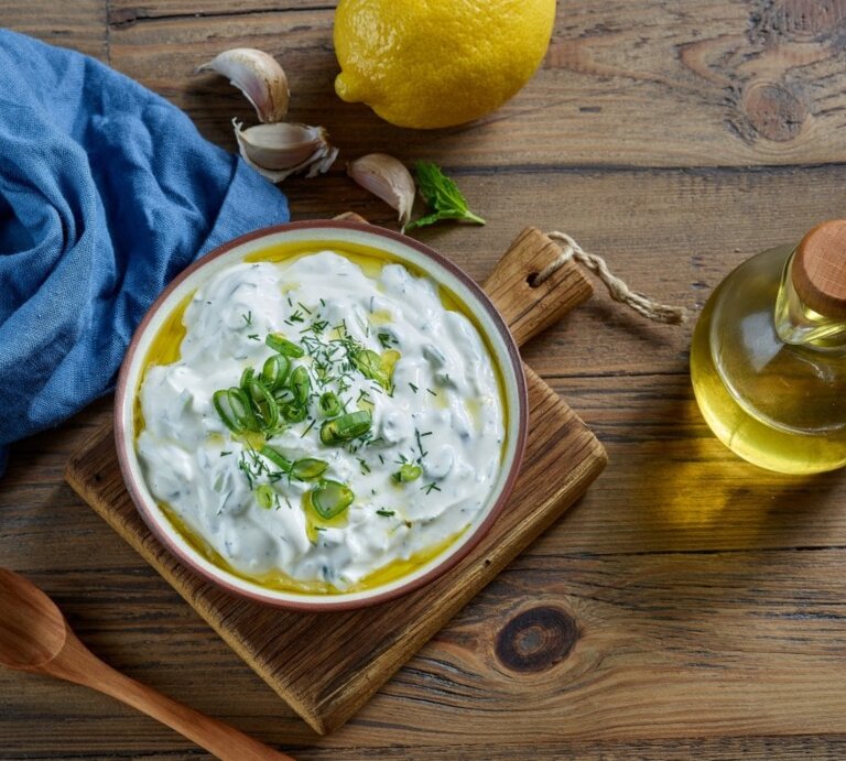 Ein farbenfrohes Bild zeigt eine Schale Çaçık, eine türkische Joghurtspeise, garniert mit geschnittenen Frühlingszwiebeln und Dill, auf einem Holzbrett. Neben der Schale befinden sich eine Zitrone, einige Knoblauchzehen, ein Bund frischer Minze und eine kleine Flasche Olivenöl mit einem Holzkorken. Eine blaue Stoffserviette liegt stilvoll gefaltet neben der Schale. Alle Elemente sind auf einer rustikalen Holzoberfläche arrangiert, die Wärme und Natürlichkeit ausstrahlt. | © Shutterstock / MaraZe
