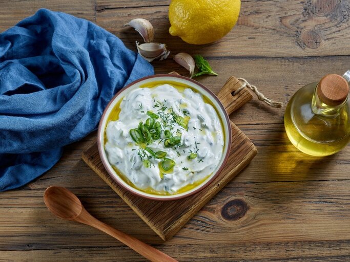 Ein farbenfrohes Bild zeigt eine Schale Çaçık, eine türkische Joghurtspeise, garniert mit geschnittenen Frühlingszwiebeln und Dill, auf einem Holzbrett. Neben der Schale befinden sich eine Zitrone, einige Knoblauchzehen, ein Bund frischer Minze und eine kleine Flasche Olivenöl mit einem Holzkorken. Eine blaue Stoffserviette liegt stilvoll gefaltet neben der Schale. Alle Elemente sind auf einer rustikalen Holzoberfläche arrangiert, die Wärme und Natürlichkeit ausstrahlt. | © Shutterstock / MaraZe