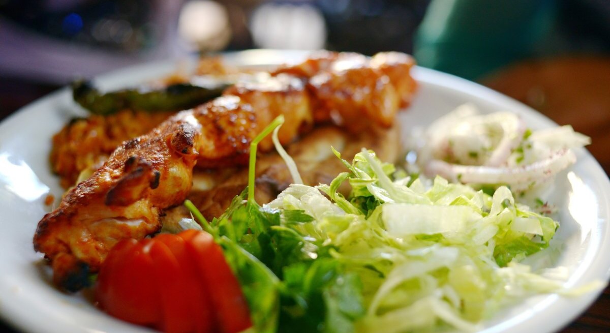 Auf einem weißen Teller sind türkische Hähnchenspieße serviert, begleitet von einem frischen, grünen Salat, einigen Tomatenscheiben und einer Portion Zwiebelringen in Joghurt-Dressing. Die Spieße sind appetitlich gebräunt und liegen im Vordergrund des Bildes, während der Salat und die Beilagen im Hintergrund verschwimmen, was auf eine geringe Tiefenschärfe hindeutet. 