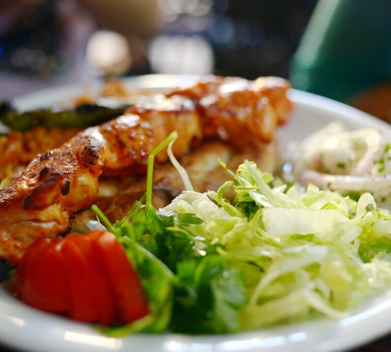 Auf einem weißen Teller sind türkische Hähnchenspieße serviert, begleitet von einem frischen, grünen Salat, einigen Tomatenscheiben und einer Portion Zwiebelringen in Joghurt-Dressing. Die Spieße sind appetitlich gebräunt und liegen im Vordergrund des Bildes, während der Salat und die Beilagen im Hintergrund verschwimmen, was auf eine geringe Tiefenschärfe hindeutet.  | © Getty Images / Tamar Mirianashvili