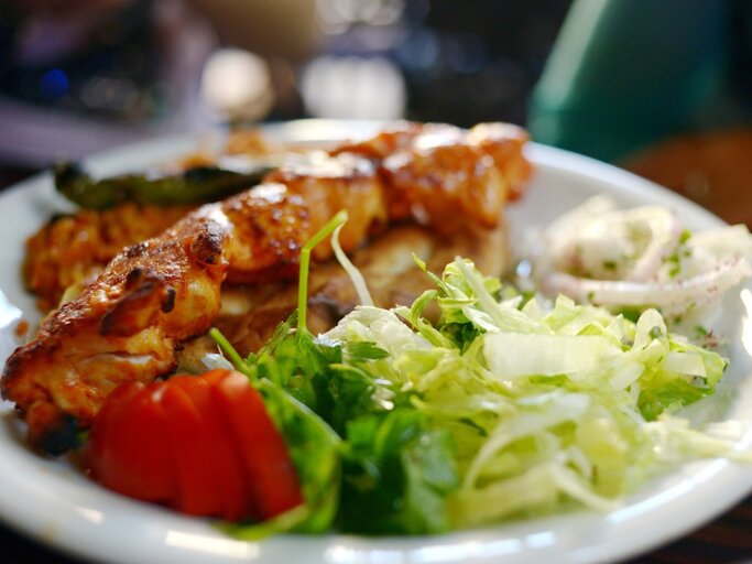 Auf einem weißen Teller sind türkische Hähnchenspieße serviert, begleitet von einem frischen, grünen Salat, einigen Tomatenscheiben und einer Portion Zwiebelringen in Joghurt-Dressing. Die Spieße sind appetitlich gebräunt und liegen im Vordergrund des Bildes, während der Salat und die Beilagen im Hintergrund verschwimmen, was auf eine geringe Tiefenschärfe hindeutet.  | © Getty Images / Tamar Mirianashvili