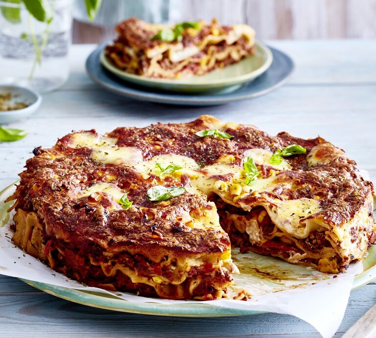 Eine angeschnittene Pizza-Lasagne auf einem gemusterten Teller auf einem blauen Untergrund. Im Hintergrund steht auf einem Teller noch ein Stück Pizza-Lasagne. | © EAT CLUB/Isabell Triemer