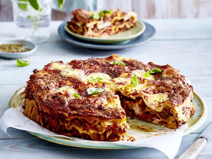 Eine angeschnittene Pizza-Lasagne auf einem gemusterten Teller auf einem blauen Untergrund. Im Hintergrund steht auf einem Teller noch ein Stück Pizza-Lasagne. | © EAT CLUB/Isabell Triemer