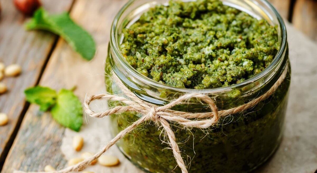 Ein Glas frisches, grünes Pesto mit rustikaler Bastschleife steht auf einem Holzbrett, umgeben von Pinienkernen und Minzblättern.