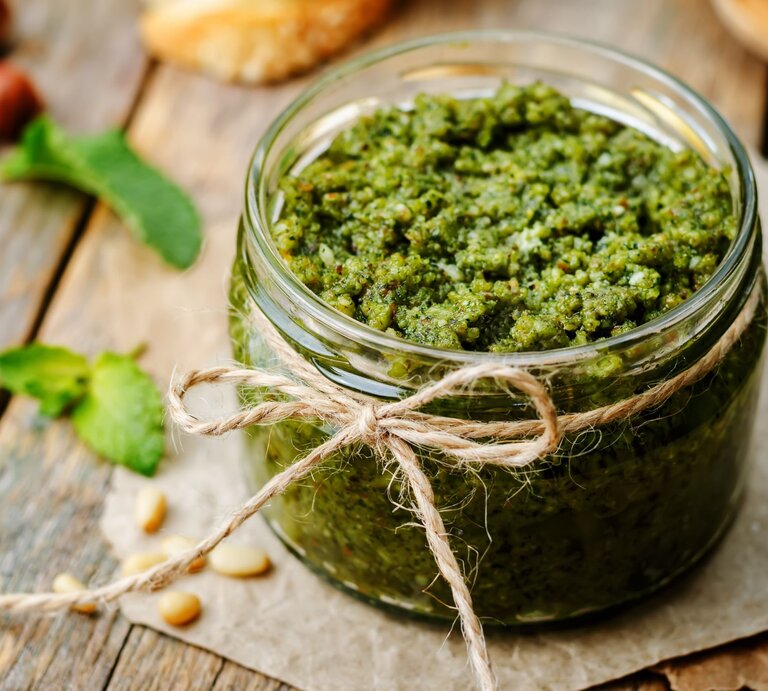 Ein Glas frisches, grünes Pesto mit rustikaler Bastschleife steht auf einem Holzbrett, umgeben von Pinienkernen und Minzblättern. | © Adobe Stock/ nata_vkusidey