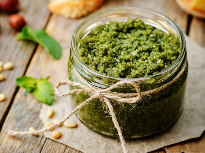 Ein Glas frisches, grünes Pesto mit rustikaler Bastschleife steht auf einem Holzbrett, umgeben von Pinienkernen und Minzblättern. | © Adobe Stock/ nata_vkusidey