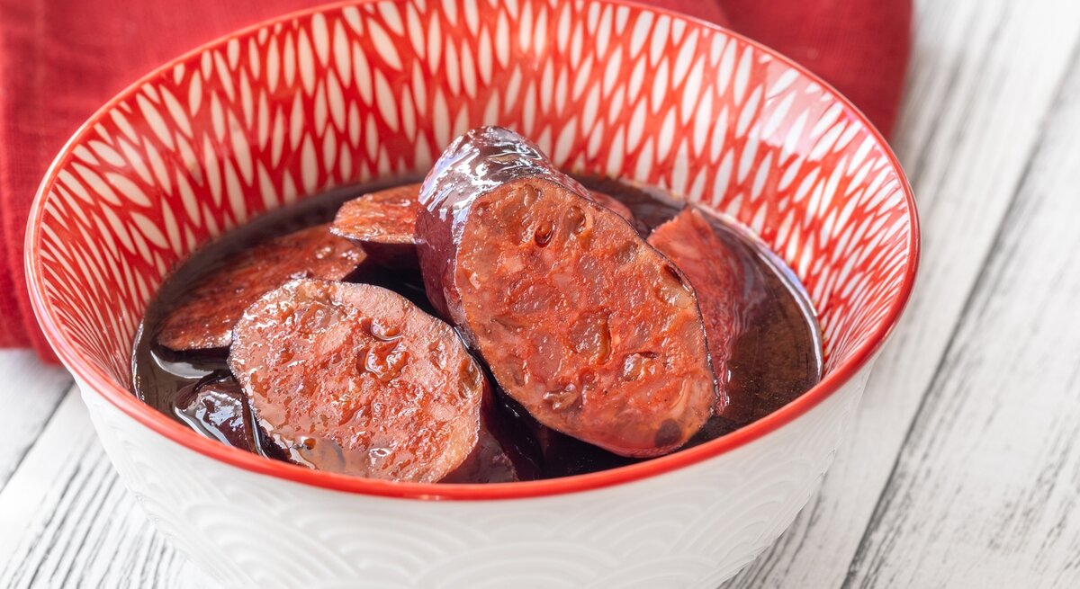 Eine weiß-rote Schale mit dem Tapasgericht auf einem hellen Holztisch. Alles von schräg oben fotografiert.