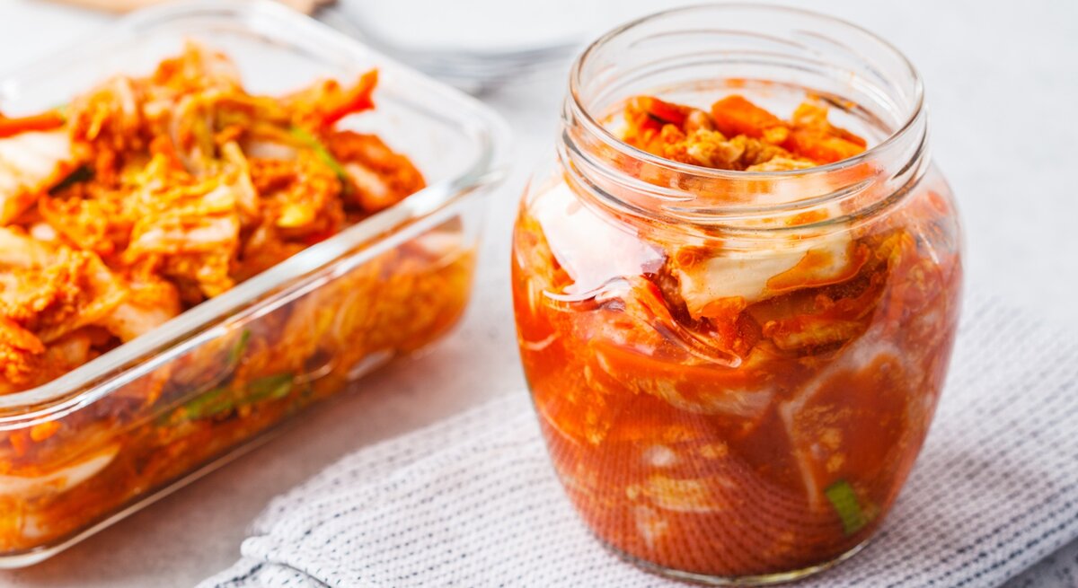 Ein Glas und eine Glas-Auflaufform mit fertigem Kimchi auf einem grauen Tisch mit weißer Tischdecke. Alles von schräg oben fotografiert.