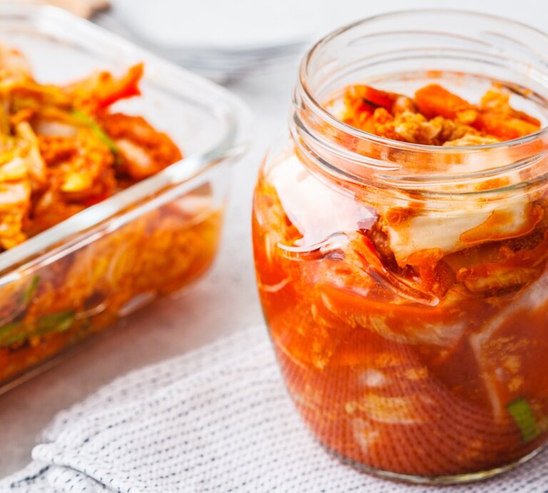 Ein Glas und eine Glas-Auflaufform mit fertigem Kimchi auf einem grauen Tisch mit weißer Tischdecke. Alles von schräg oben fotografiert. | © Shutterstock/ Nina Firsova
