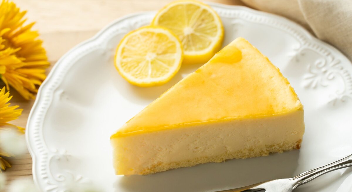 Ein Stück Zitronenkuchen mit glänzend gelber Glasur liegt auf einem verzierten weißen Teller, daneben Zitronenscheiben und gelbe Blüten.