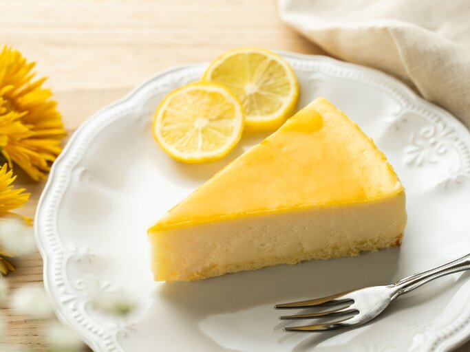 Ein Stück Zitronenkuchen mit glänzend gelber Glasur liegt auf einem verzierten weißen Teller, daneben Zitronenscheiben und gelbe Blüten. | © Adobe Stock/ shironagasukujira