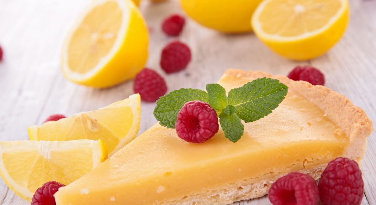 Zitronenkuchen-Stück im Vordergrund, belegt mit Himbeeren und im Hintergrund aufgeschnittene Zitronen