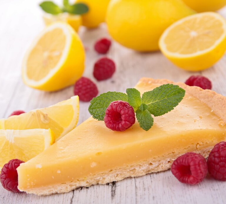 Zitronenkuchen-Stück im Vordergrund, belegt mit Himbeeren und im Hintergrund aufgeschnittene Zitronen | © gettyimages/ margouillatphotos