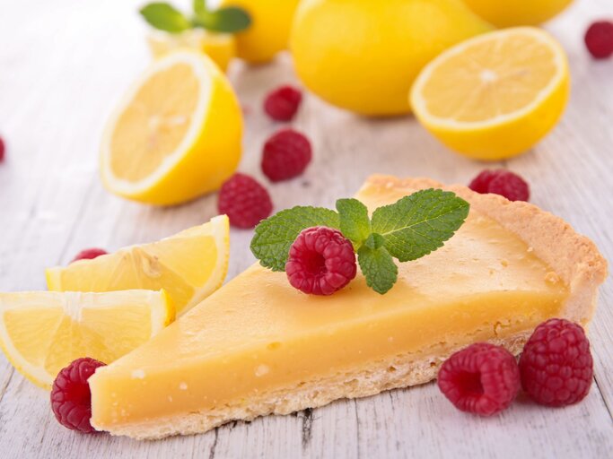 Zitronenkuchen-Stück im Vordergrund, belegt mit Himbeeren und im Hintergrund aufgeschnittene Zitronen | © gettyimages/ margouillatphotos