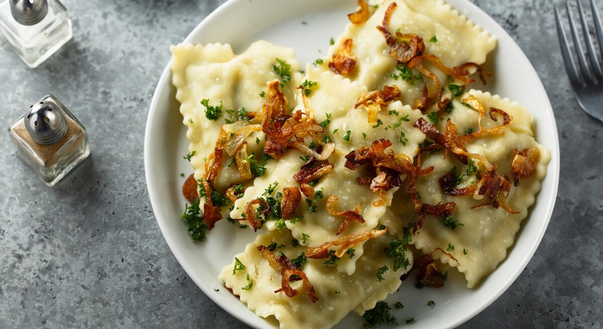 Mehrere Maultaschen mit Kräutern und Röstzwiebeln garniert auf weißem Teller und grauem Untergrund.