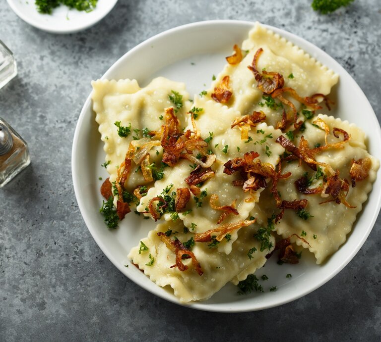 Mehrere Maultaschen mit Kräutern und Röstzwiebeln garniert auf weißem Teller und grauem Untergrund. | © Shutterstock/ MariaKovaleva
