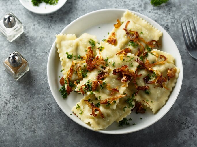 Mehrere Maultaschen mit Kräutern und Röstzwiebeln garniert auf weißem Teller und grauem Untergrund. | © Shutterstock/ MariaKovaleva