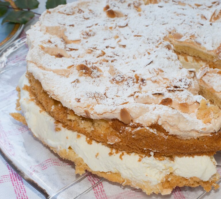 Die Sahnetorte mit Baiserschicht und mandeln auf einem Tisch mit rot-weiß-karierter Tischdecke | © Adobe Stock/ vschlichting
