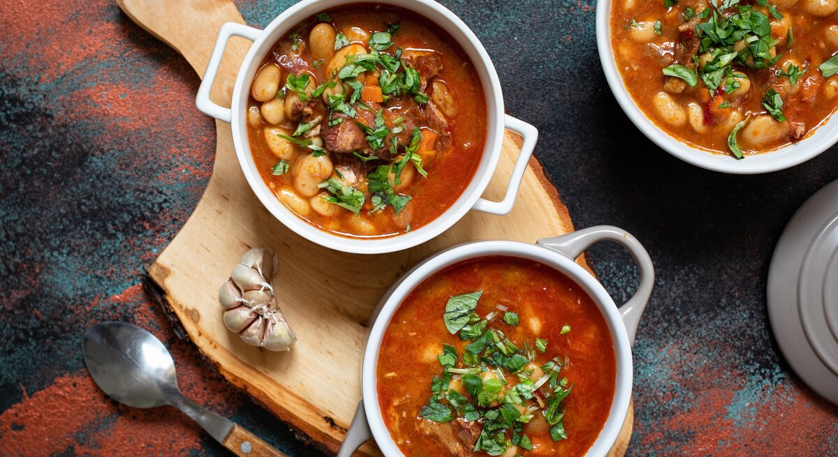Serbische Bohnensuppe in drei runden Schalen aus Porzellan / Holzbrett / frischer Knoblauch / Suppenlöffel / Draufsicht